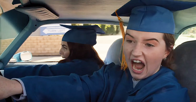 Beanie Feldstein and Kaitlyn Dever in their graduation caps and gowns, driving insanely.