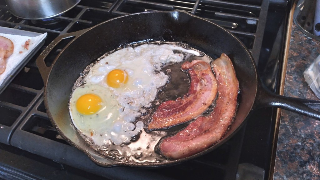 Two eggs added next to the slices of bacon, frying in the iron skillet