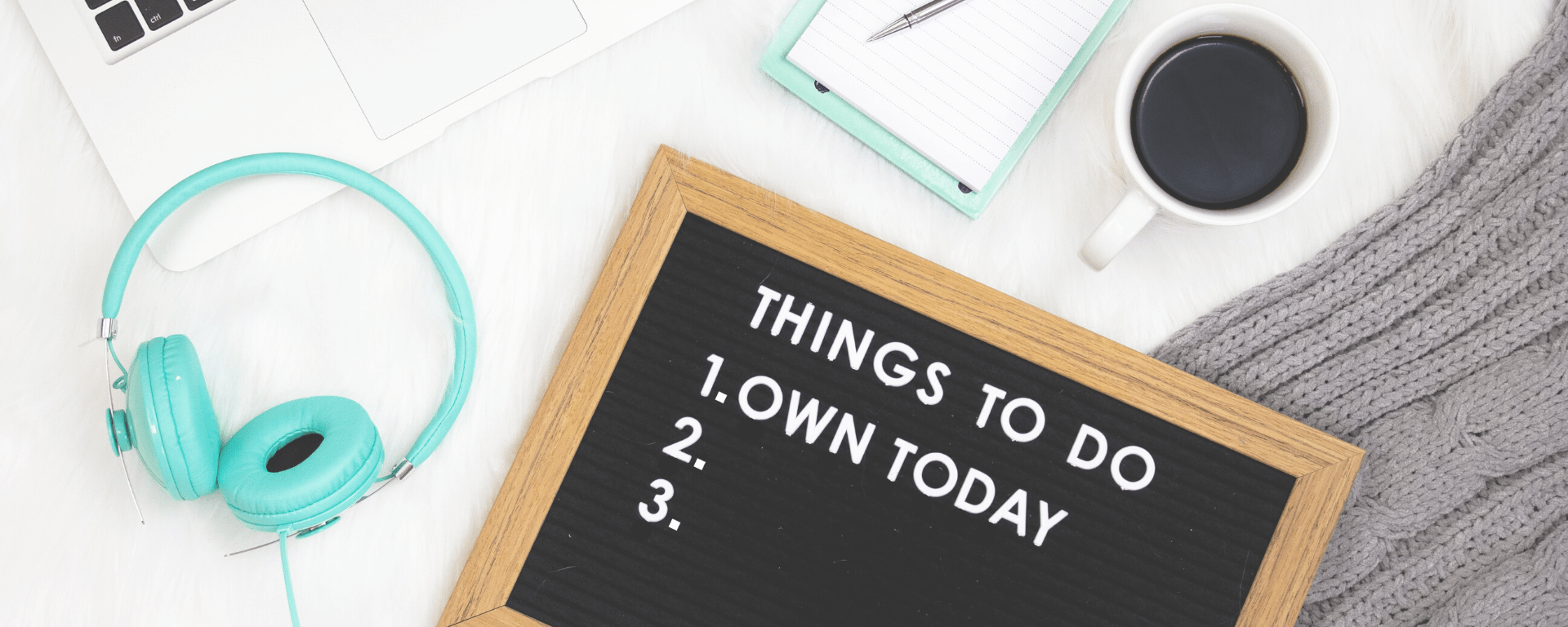 Teal-colored headphones with a letter board and notepad and cup of coffee next to a MacBook laptop